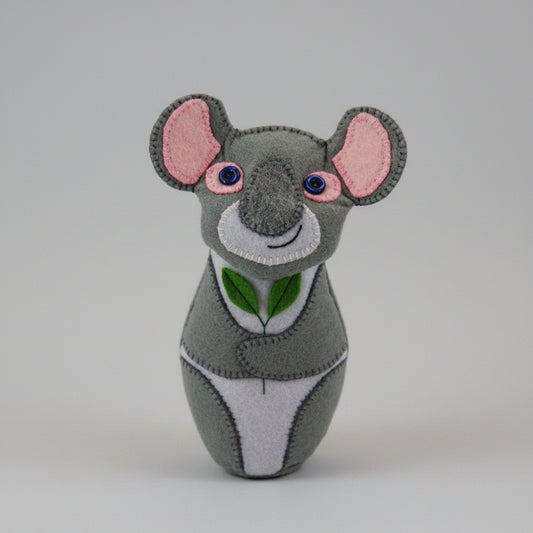 Rankų darbo pilka su baltomis ir rožinėmis detalėmis koala, laikanti žalią eukalipto šakelę